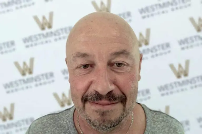 Middle-aged man with hair regrowth, sporting a shaved head and goatee, wearing a gray t-shirt, smiling at the camera. The background features the logo of Westminster repeatedly.