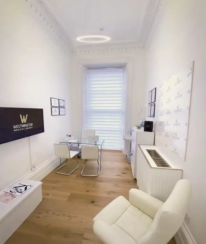 Interior of a modern, minimalistic office with white walls, a small desk with a computer, four chairs, and promotional material on female hair loss on the walls. A large window lets in natural light