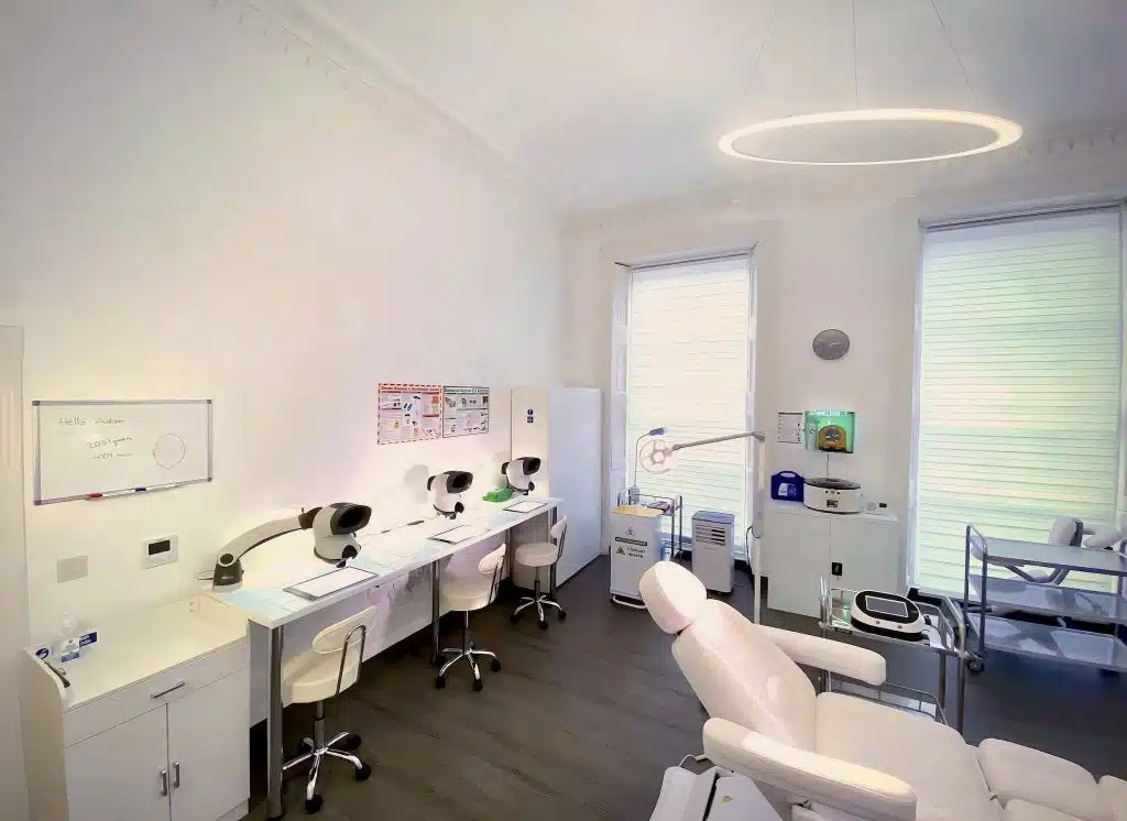 A modern dental clinic room with two dental chairs, overhead lights, a whiteboard, and a desk with computers focused on treatments including hair regrowth. The room has white walls and closed blinds on windows