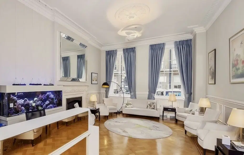 Elegant living room with a large aquarium, white fireplace, blue curtains, and plush seating under a decorative ceiling and chandelier, overlooking city views.