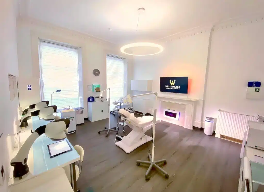 A modern dental clinic room with two dental chairs, overhead lights, and equipment. A monitor displays a logo, and the room features bright, clean décor and wooden flooring.