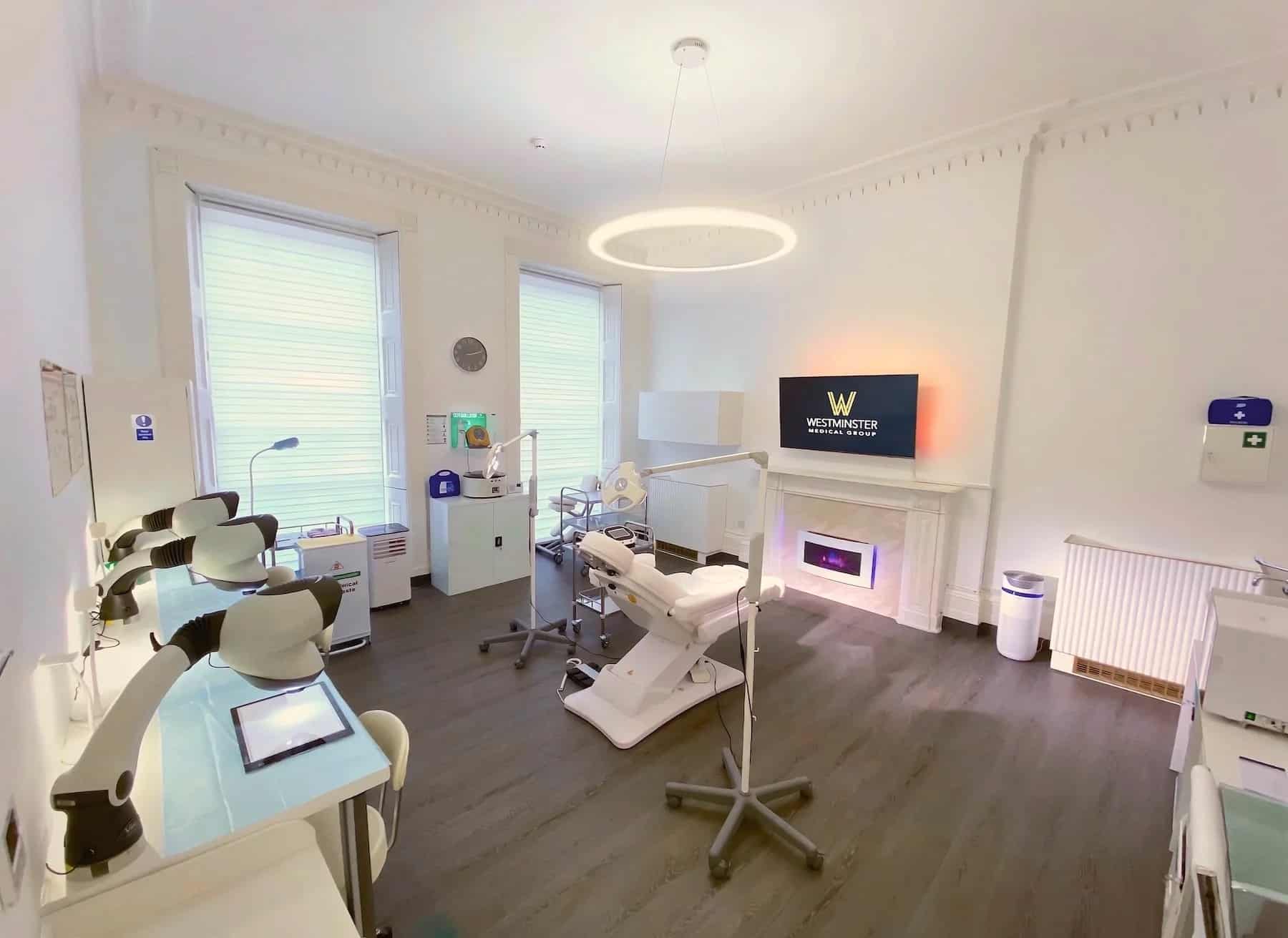 Modern dental clinic interior with two dental chairs, a TV displaying a clinic logo specialized in treatments for female hair loss, and clean, bright decor with white walls and wooden floors.