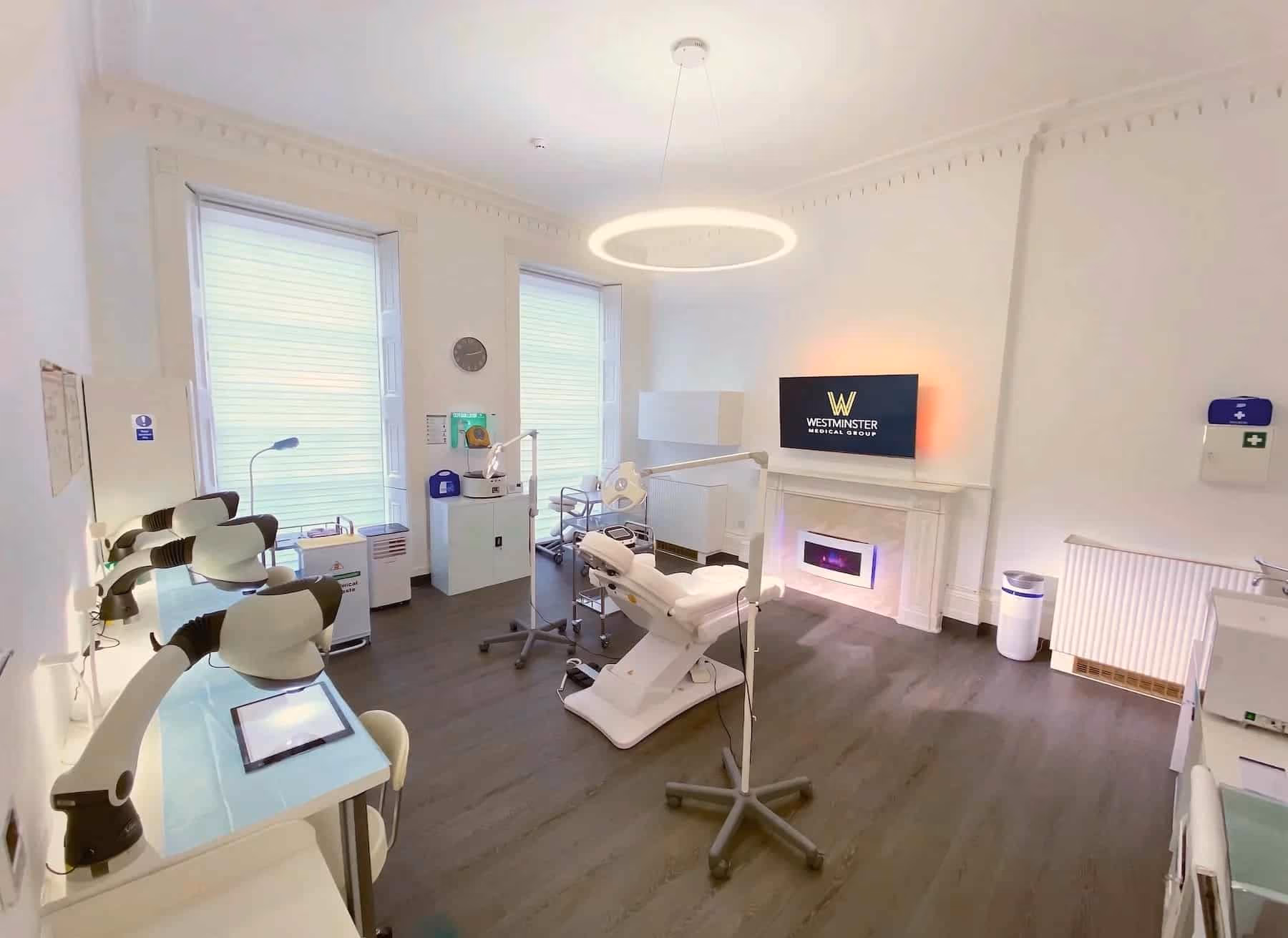 Modern dental clinic interior with two dental chairs, a TV displaying a clinic logo specialized in treatments for female hair loss, and clean, bright decor with white walls and wooden floors.