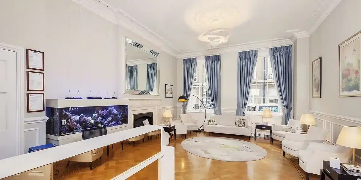 Elegant, spacious living room with white walls, a large aquarium, ornate fireplace, plush sofas, and long curtains on windows providing natural light. Decor includes a ceiling rosette and various artworks
