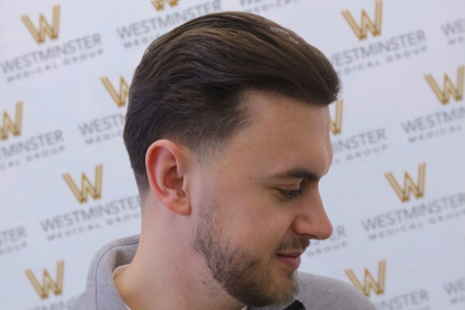 Profile view of a young man with styled hair, smiling slightly, in front of a backdrop with the "Westminster" logo repeated.